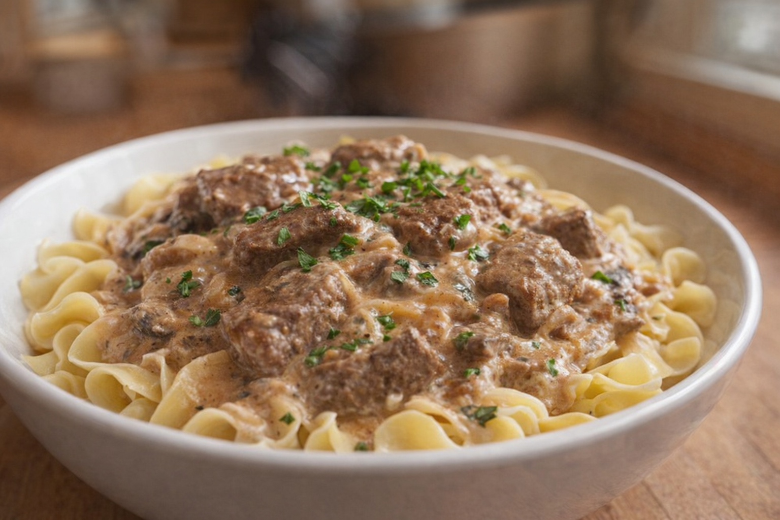 A close-up shot of Beef Stroganoff served over egg noodles in a white bowl, garnished with fresh parsley. The sauce is creamy and rich, and the beef is tender. Warm, inviting lighting.
